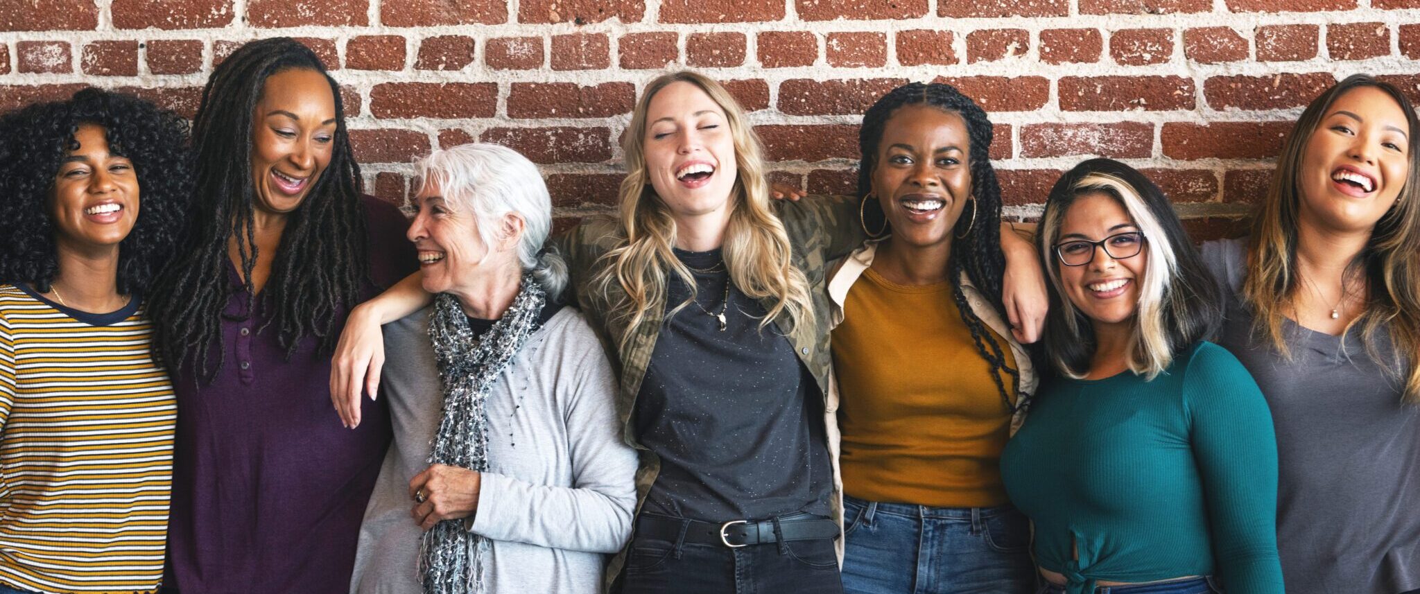 Diverse group of women smiling and standing together against a brick wall.